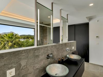 Salle de bain contemporaine avec double vasque en verre, robinetterie murale et mur carrelé texturé gris. Les miroirs éclairés reflètent la douche et la fenêtre panoramique sur les montagnes.