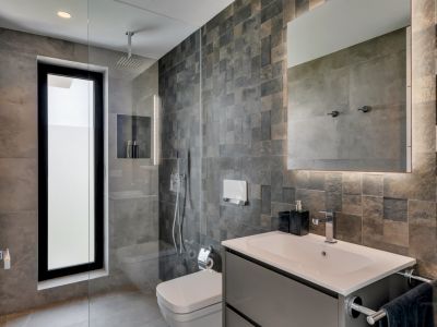 Salle de bain moderne avec carrelage mural texturé foncé, douche vitrée à l'italienne et grande fenêtre verticale. Un miroir rétroéclairé éclaire la vasque et son meuble gris.