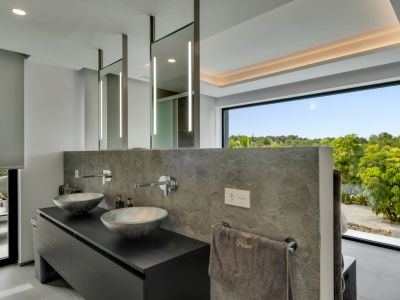 Salle de bain moderne avec double vasque, lavabos design et robinetterie murale. Miroirs éclairés et grande fenêtre offrant une vue sur la verdure luxuriante.