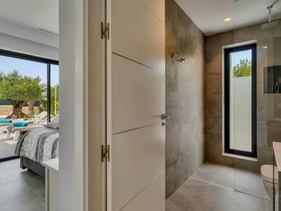 Salle de bain moderne avec douche à l'italienne, murs carrelés gris et fenêtre dépolie, attenante à une chambre avec accès direct au jardin et un olivier.