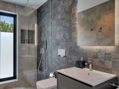 Salle de bain moderne avec douche à l'italienne vitrée et pommeau de pluie. Carrelage mural texturé gris foncé, fenêtre dépolie et miroir rétroéclairé au-dessus d'une vasque épurée.