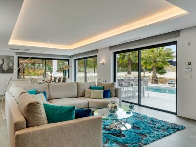Salon contemporain avec canapé d'angle, tapis turquoise et grandes baies vitrées offrant un accès direct à la piscine et aux palmiers.