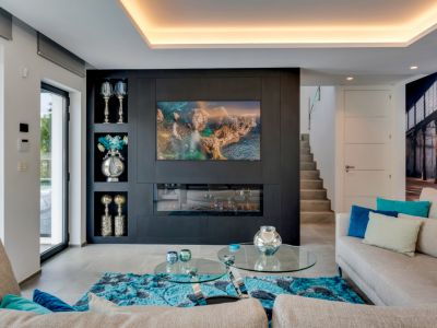 Salon moderne doté d'un mur d'accent sombre avec télévision et cheminée électrique intégrées, éclairage indirect et grandes portes vitrées vers l'extérieur.
