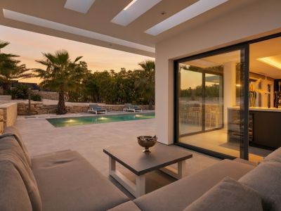 Terrasse couverte avec canapé modulaire et table basse, offrant une vue sur la piscine éclairée, les palmiers et le ciel au coucher du soleil. De grandes baies vitrées mènent à l'intérieur.