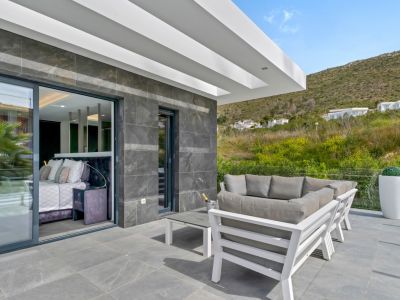 Terrasse extérieure carrelée de gris avec un mobilier de salon blanc moderne. De grandes baies vitrées s'ouvrent sur une chambre intérieure, offrant une vue sur la colline verdoyante.