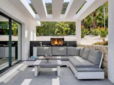 Terrasse extérieure couverte moderne avec cuisine intégrée et barbecue allumé, dotée d'un canapé d'angle et d'une table basse sur carrelage gris, face à un jardin paysager.