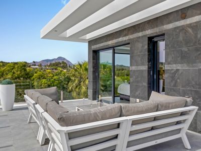 Terrasse extérieure moderne avec canapé gris en L, murs carrelés sombres et balustrade en verre. Vues sur les montagnes et la verdure, avec accès direct à une pièce intérieure.