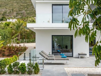 Terrasse extérieure moderne avec carrelage gris et chaises longues contemporaines, dotée d'une balustrade en verre offrant des vues sur un jardin méditerranéen luxuriant et les montagnes.