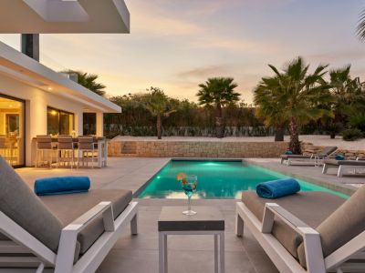 Terrasse extérieure moderne avec piscine éclairée au crépuscule, entourée de chaises longues contemporaines et un bar extérieur visible à travers des baies vitrées.