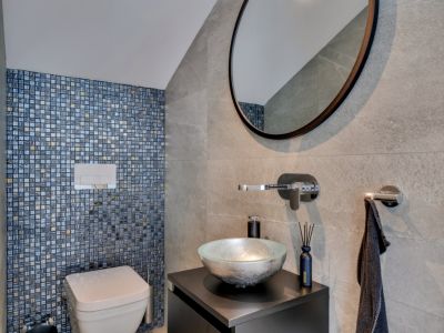 Toilettes d'invités modernes avec mur d'accent en mosaïque bleue et dorée, WC suspendu et lavabo en verre argenté sur meuble sombre.