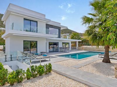 Villa blanche moderne avec balcons vitrés et piscine rectangulaire. L'extérieur présente une terrasse carrelée grise, aménagement en gravier, salle à manger extérieure et transats, sur fond montagneux.