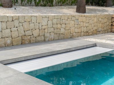 Volet roulant automatique blanc protégeant la piscine, entouré de dalles grises et d'un mur de soutènement en pierre naturelle, avec un jardin de gravier.