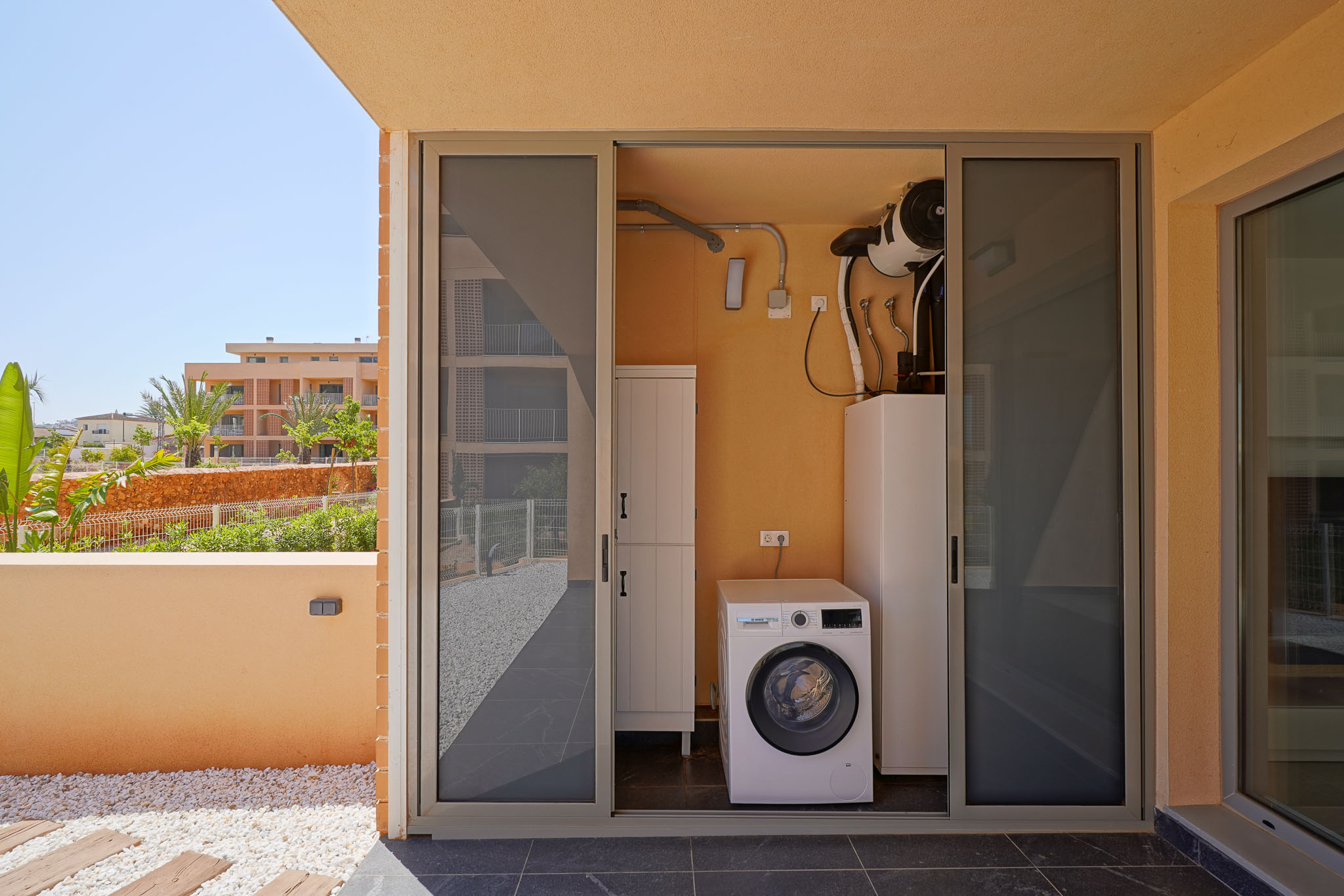 Buanderie extérieure avec machine à laver Bosch et système aérothermique, accessible depuis une terrasse carrelée avec galets décoratifs et vue sur jardin.