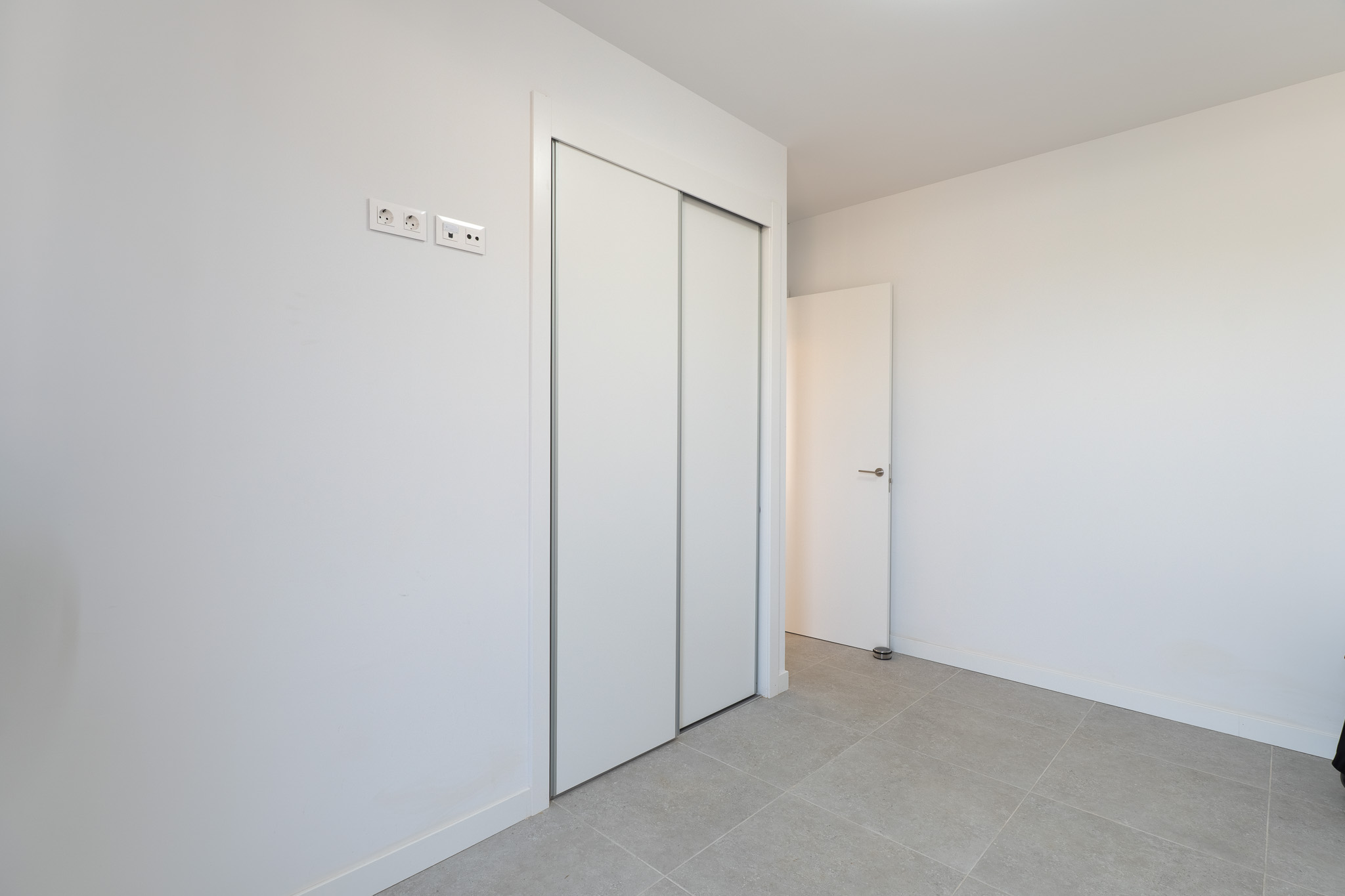 Chambre avec un grand placard intégré à portes coulissantes blanches et un sol carrelé gris clair. Des prises électriques et de données sont installées.