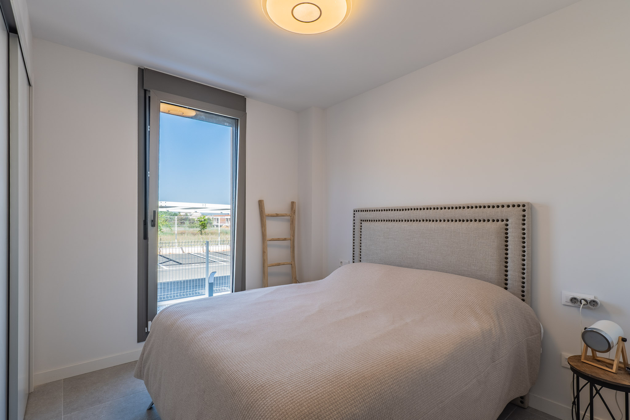Chambre lumineuse avec lit, tête de lit en tissu clouté et armoire intégrée. Grande fenêtre offrant un accès extérieur et un sol carrelé gris.
