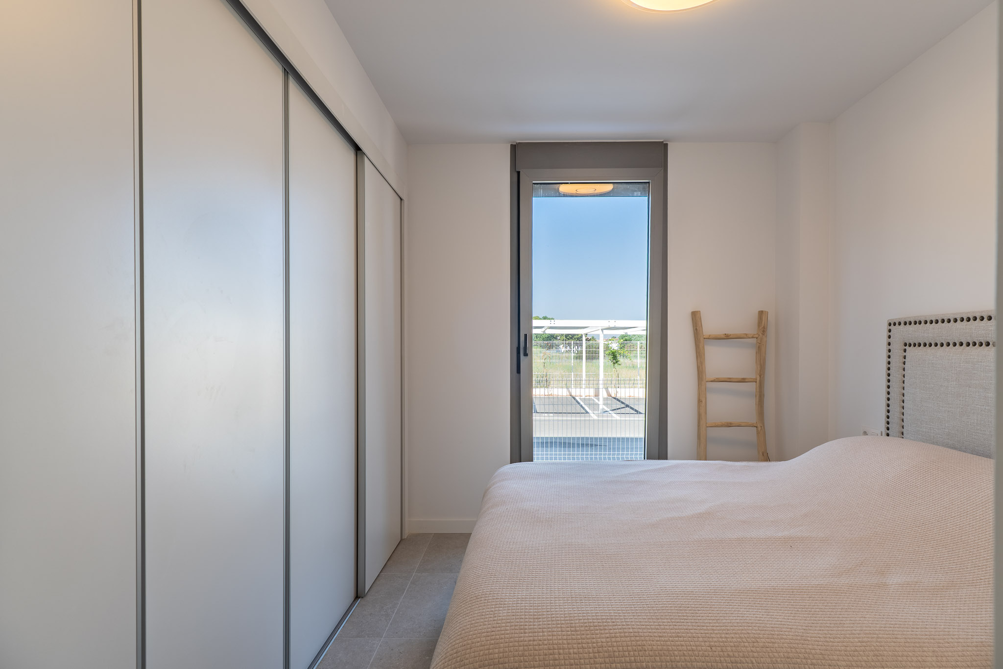 Chambre lumineuse avec placard intégré à portes coulissantes blanches, sol carrelé et accès direct à l'extérieur par une porte vitrée. Lit avec tête de lit capitonnée.