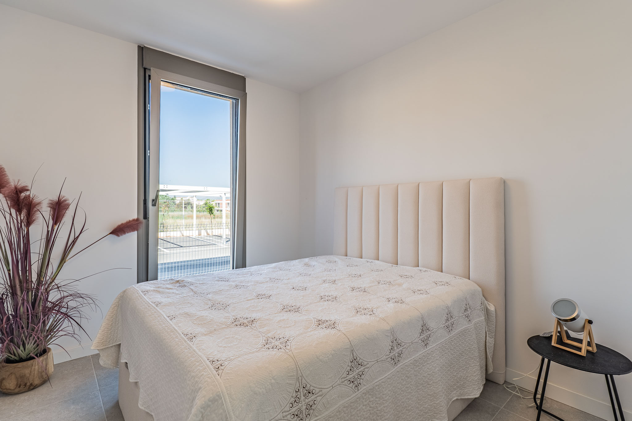 Chambre lumineuse avec un lit et une tête de lit rembourrée beige. Grande fenêtre du sol au plafond offrant une vue extérieure. Sol carrelé gris et plante décorative.