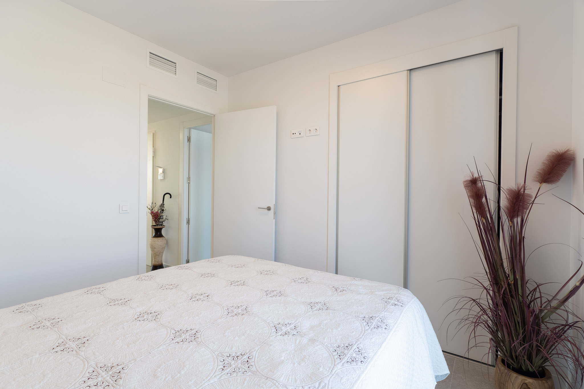 Chambre lumineuse avec un lit orné d'un couvre-lit blanc brodé. Elle dispose d'une armoire encastrée à portes coulissantes et de bouches de climatisation au plafond. Une plante décorative agrémente l'espace.