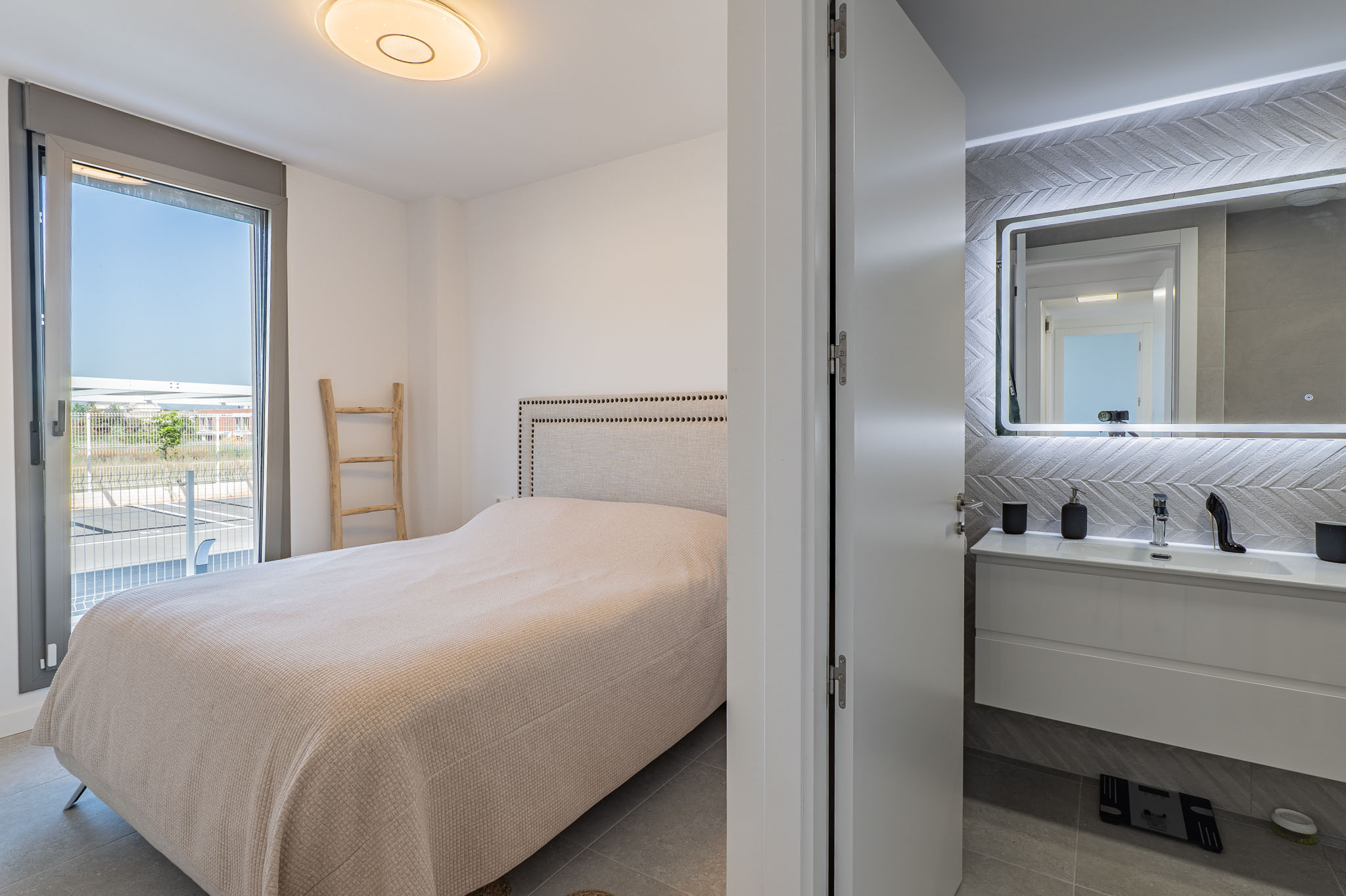 Chambre principale avec lit à tête de lit rembourrée et accès direct à une terrasse extérieure. Une salle de bain attenante avec meuble-lavabo suspendu et miroir rétroéclairé est visible.