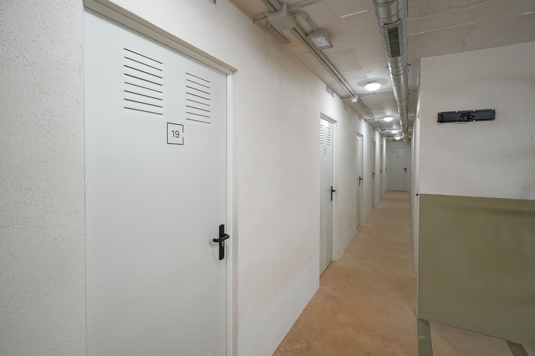 Couloir de stockage avec des portes blanches à persiennes, numérotées, et poignées noires. Le plafond révèle des conduits apparents et un éclairage encastré, au-dessus d'un sol en béton.