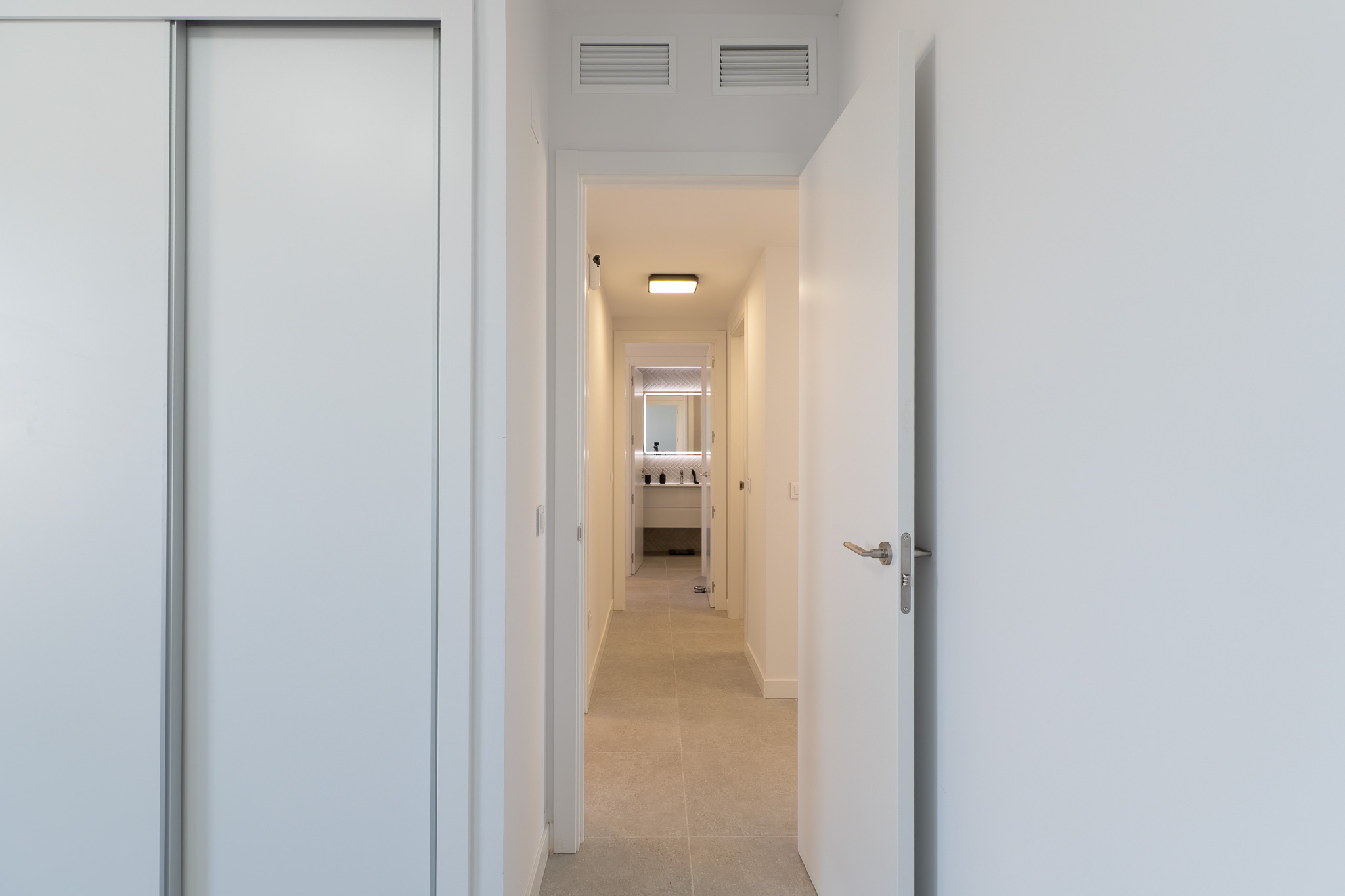 Couloir lumineux avec placard coulissant blanc et porte ouverte. Le carrelage clair mène à une salle de bain moderne avec miroir rétroéclairé et meuble-lavabo contemporain. Grilles de ventilation au plafond.