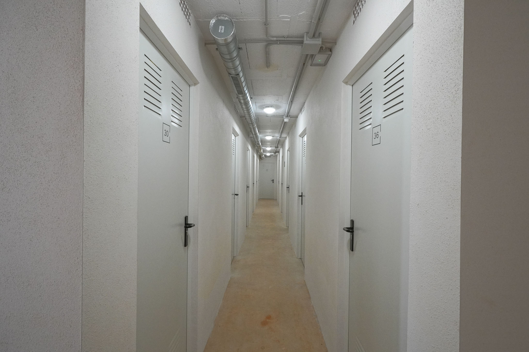 Couloir utilitaire vers des unités de stockage individuelles, avec portes blanches numérotées et fentes d'aération. Plafond en béton apparent avec conduits industriels et éclairage encastré, murs texturés.