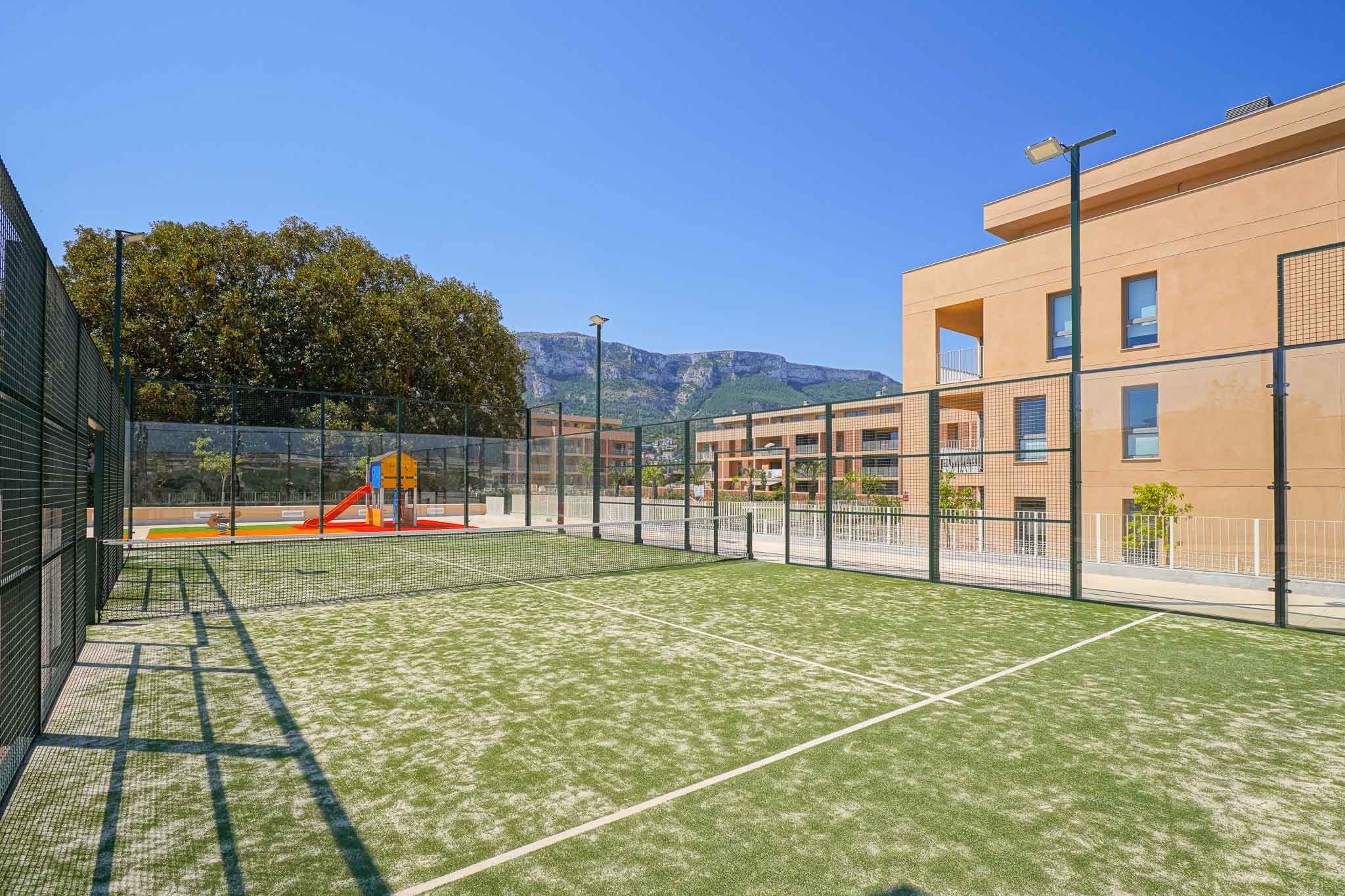 Court de padel en gazon synthétique vert, clôturé et éclairé, jouxtant une aire de jeux pour enfants et des bâtiments résidentiels modernes avec vue sur les montagnes.