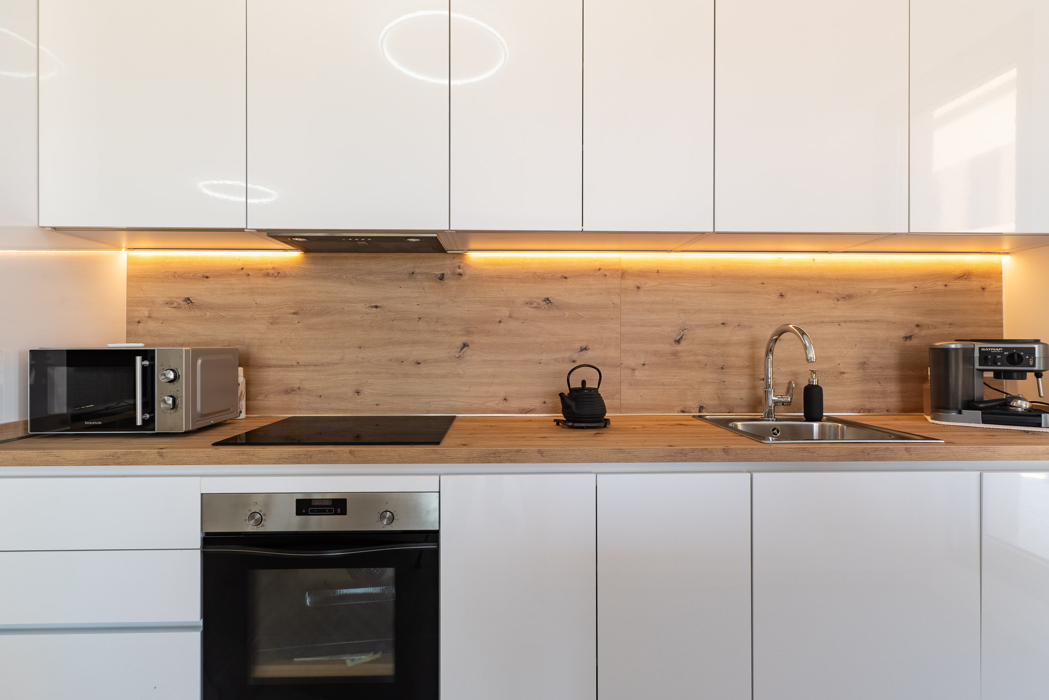 Cuisine moderne avec des armoires blanches laquées, un plan de travail et une crédence effet bois, et un éclairage LED sous les meubles hauts. Four encastré, plaque à induction et micro-ondes.