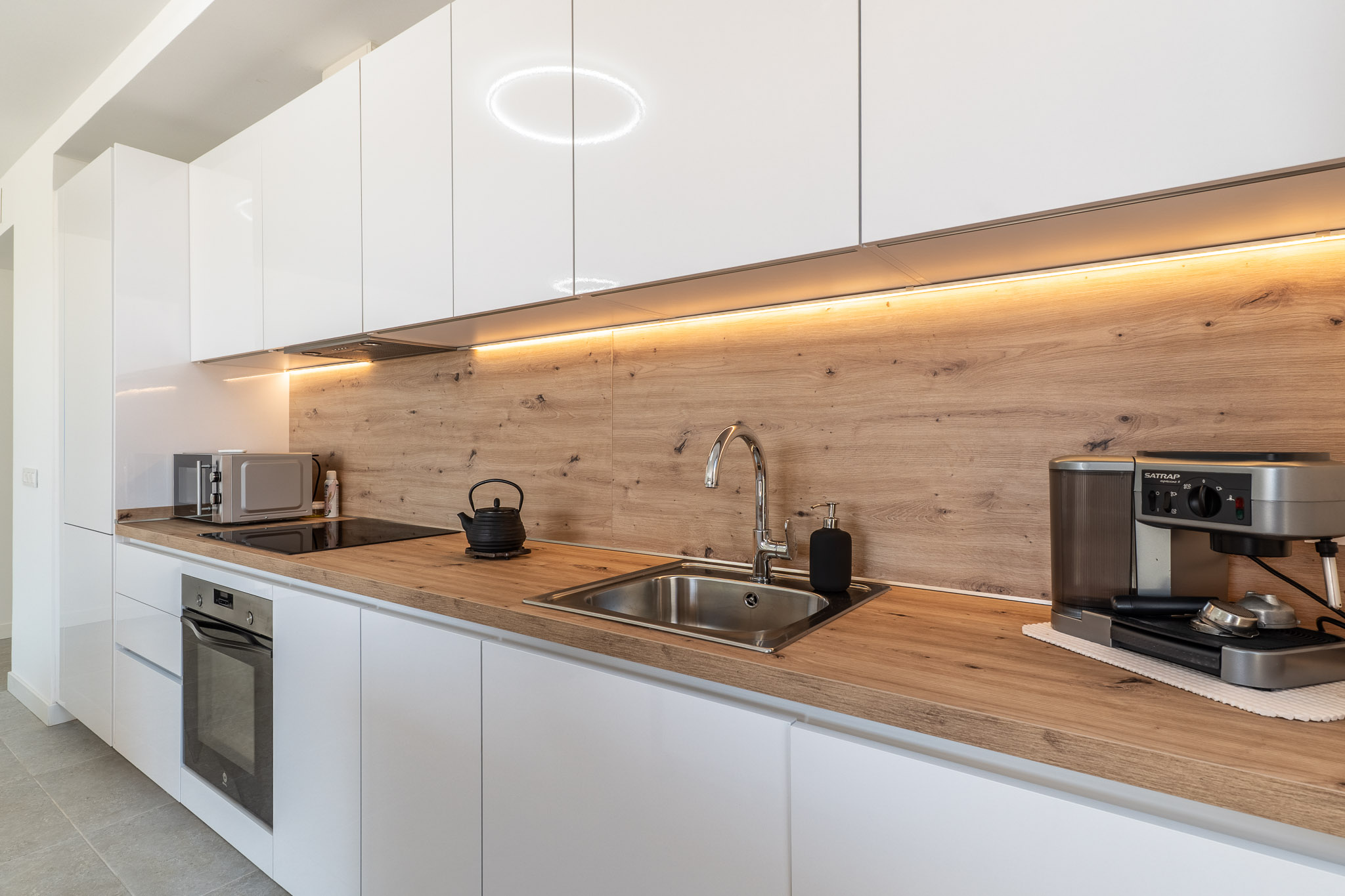 Cuisine moderne avec meubles blancs laqués, plan de travail et crédence aspect bois clair. Elle intègre un four, une plaque à induction et un évier en inox, soulignée par un éclairage LED sous les placards.