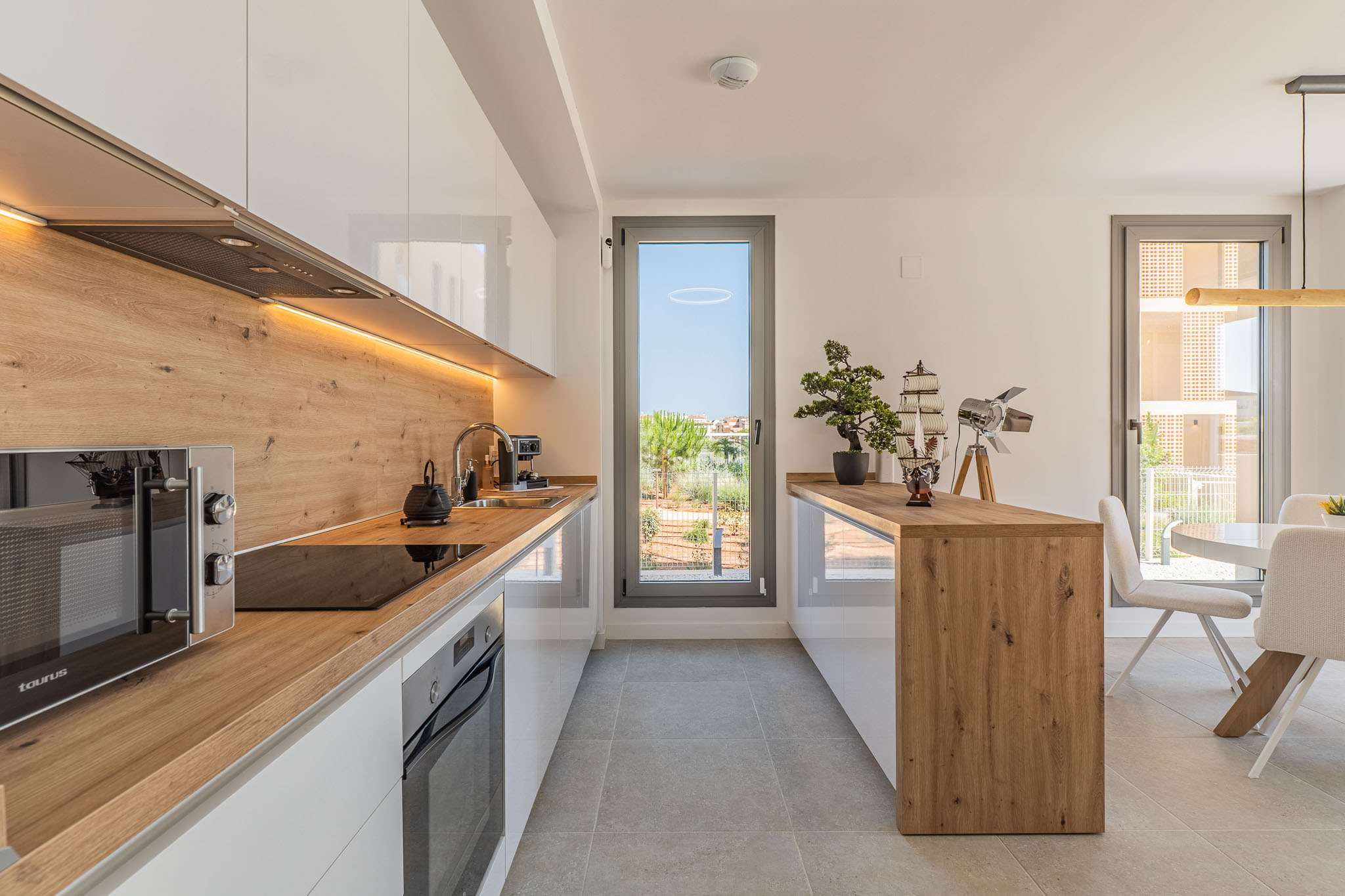 Cuisine ouverte moderne avec des armoires blanches laquées, des plans de travail et crédence effet bois, et un éclairage LED sous les meubles. Équipée d'appareils intégrés et ouvrant sur l'extérieur.