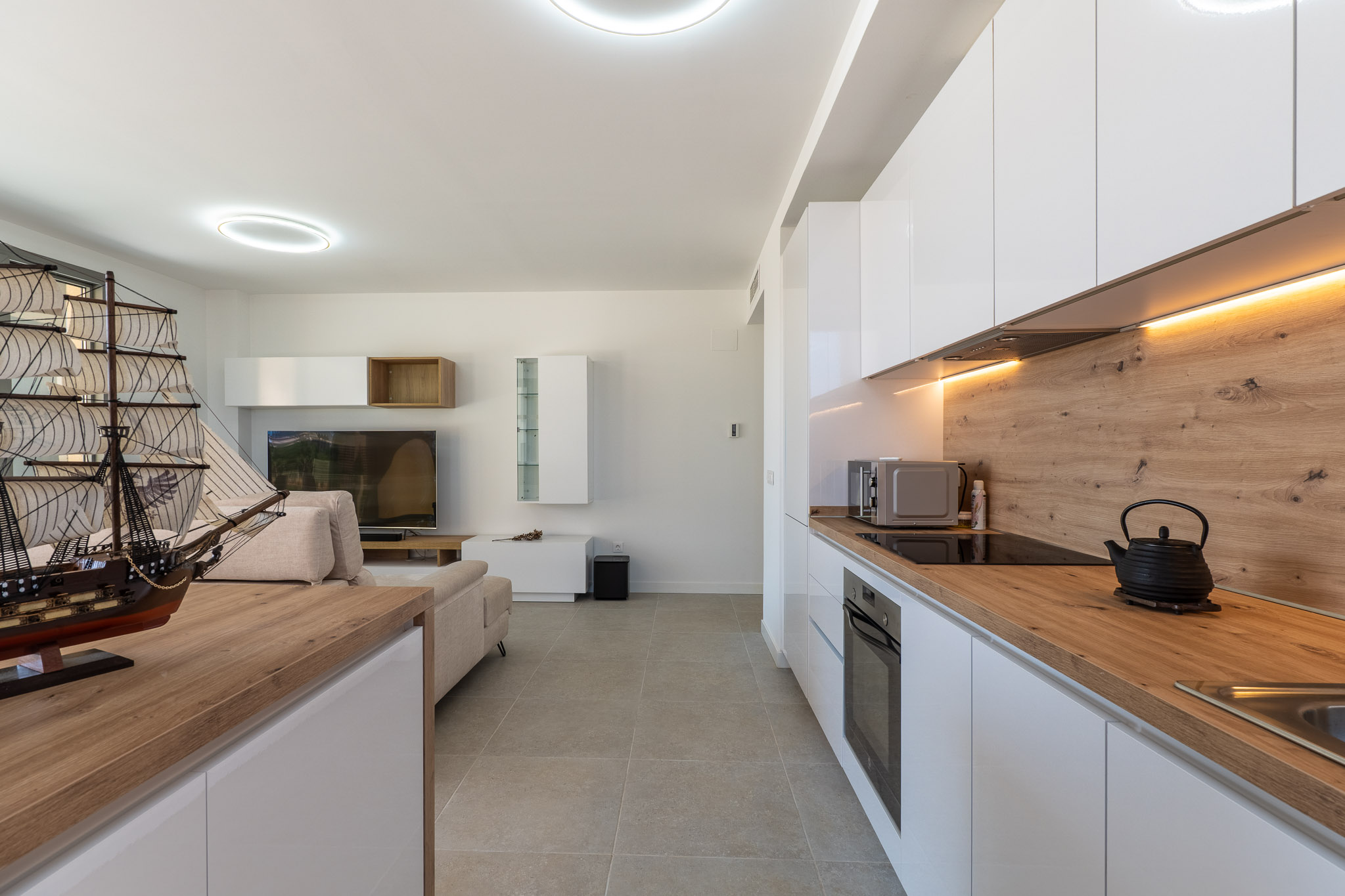 Espace de vie ouvert intégrant un salon et une cuisine moderne. La cuisine présente des armoires laquées blanches, des plans de travail effet bois clair, des appareils intégrés et un éclairage LED sous les meubles.