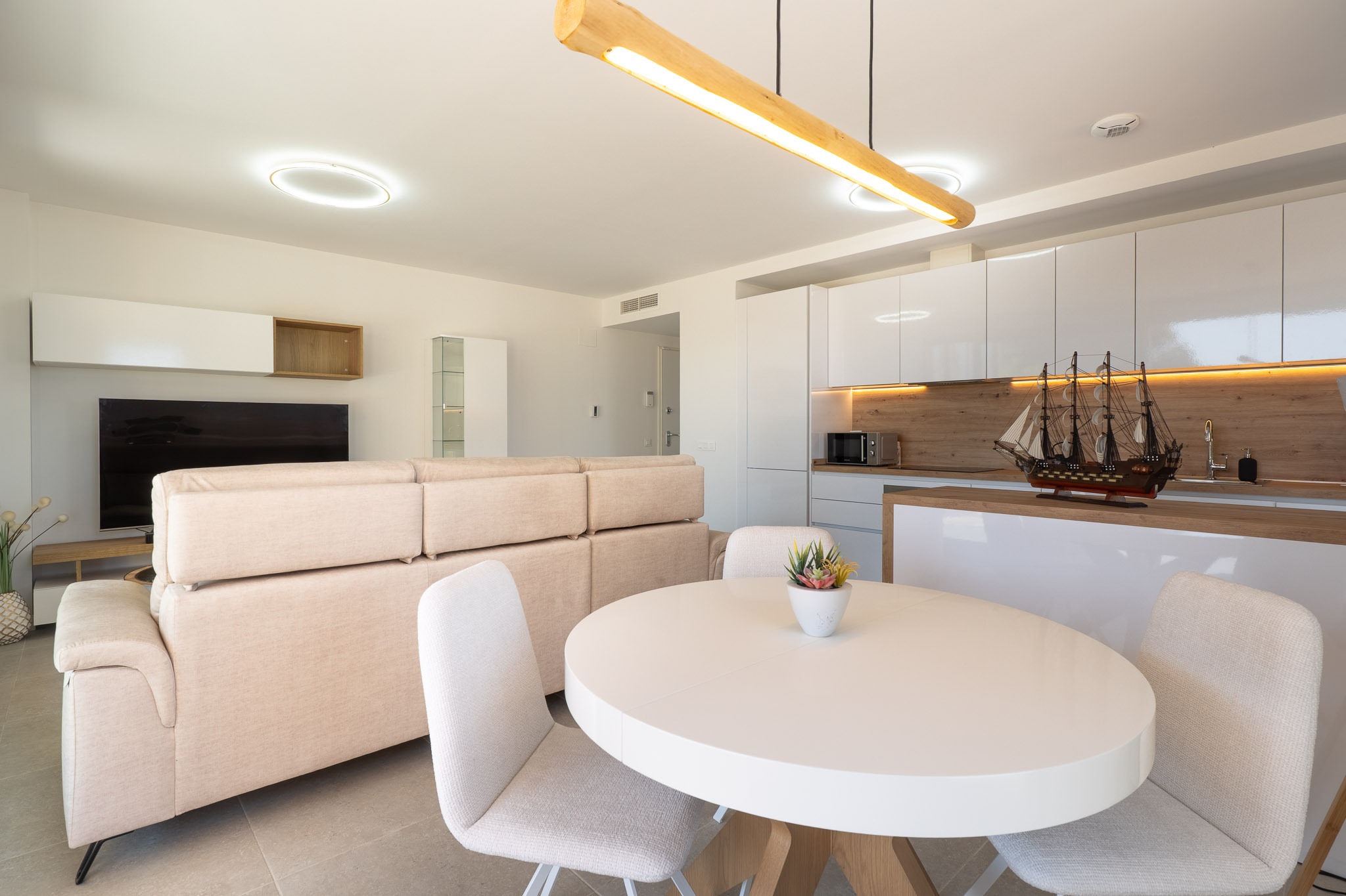 Espace salon-salle à manger ouvert avec cuisine intégrée. Il présente un canapé clair, une table ovale blanche et une cuisine moderne avec des meubles laqués blancs et des accents boisés.
