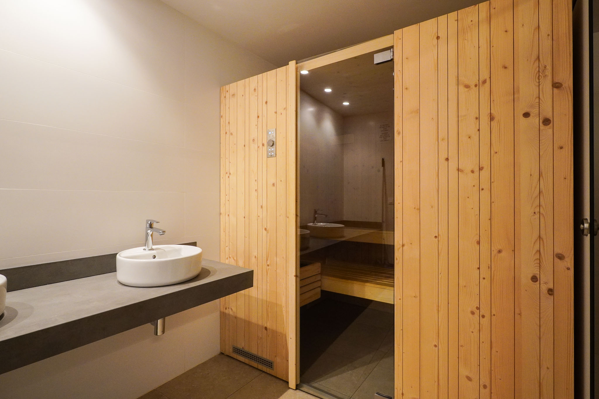 Espace vestiaire avec un lavabo vasque design sur meuble flottant et l'entrée du sauna, dotée d'une porte en bois à lattes et d'un panneau vitré.