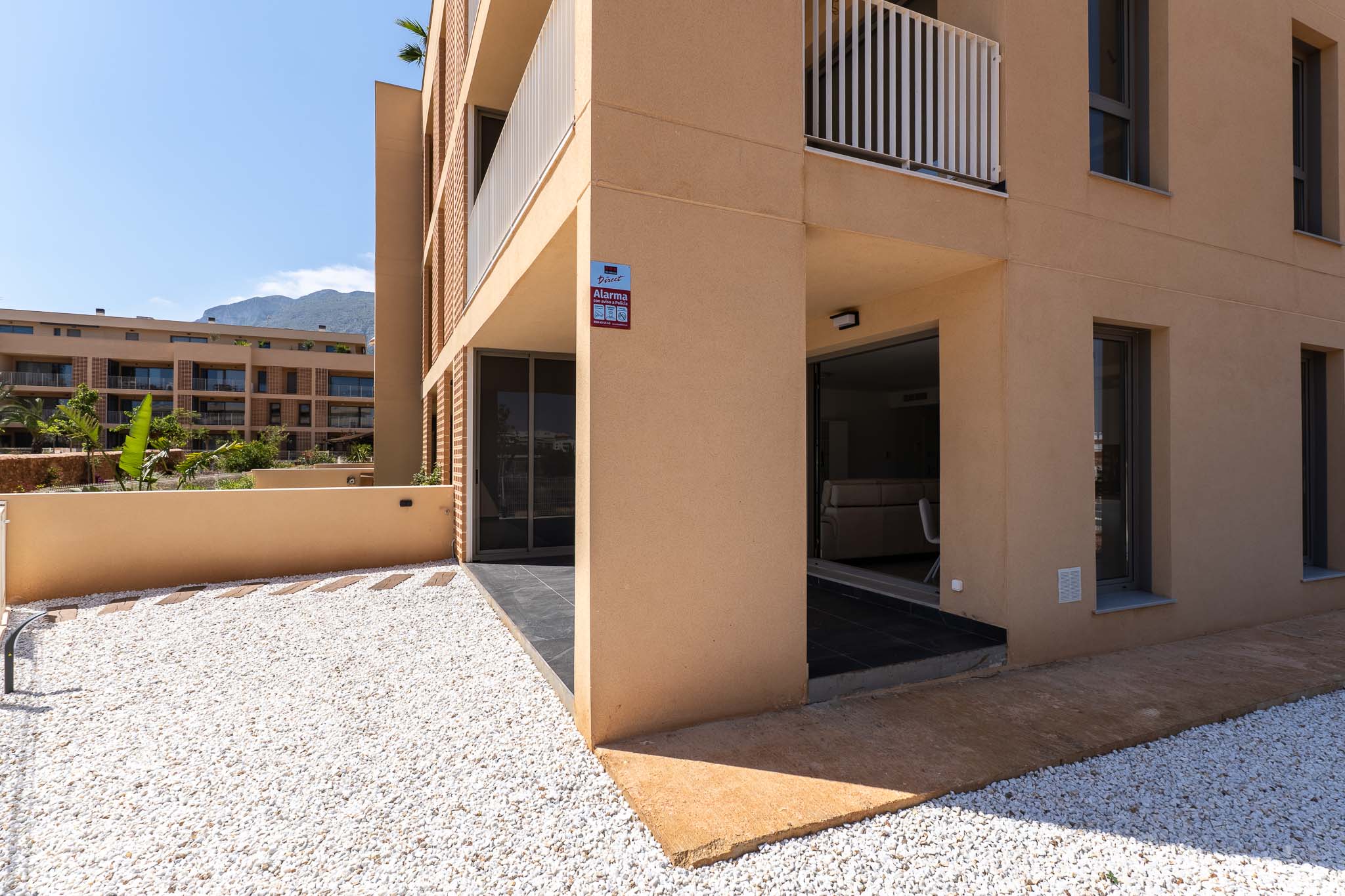Extérieur de rez-de-chaussée avec façade ocre moderne. Il comprend un jardin privé en gravier blanc et une terrasse couverte avec accès direct à l'intérieur, vue sur la montagne.