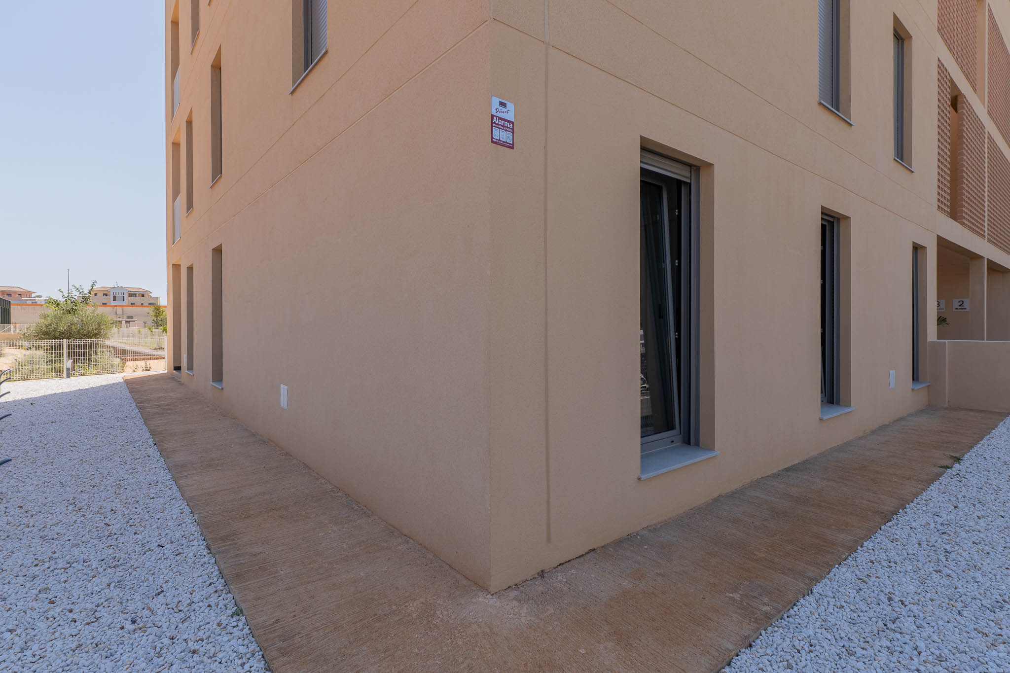 Extérieur d'un bâtiment moderne avec façade en stuc beige clair et fenêtres à cadres sombres. Le sol combine un chemin en béton texturé et un aménagement paysager de gravier blanc, avec alarme visible.