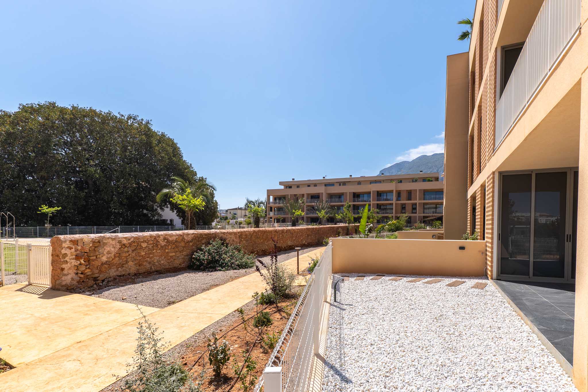 Jardin privé en rez-de-chaussée avec gravier blanc et dalles, délimité par une clôture basse et un mur en pierre. Vues sur les bâtiments modernes et les montagnes lointaines.
