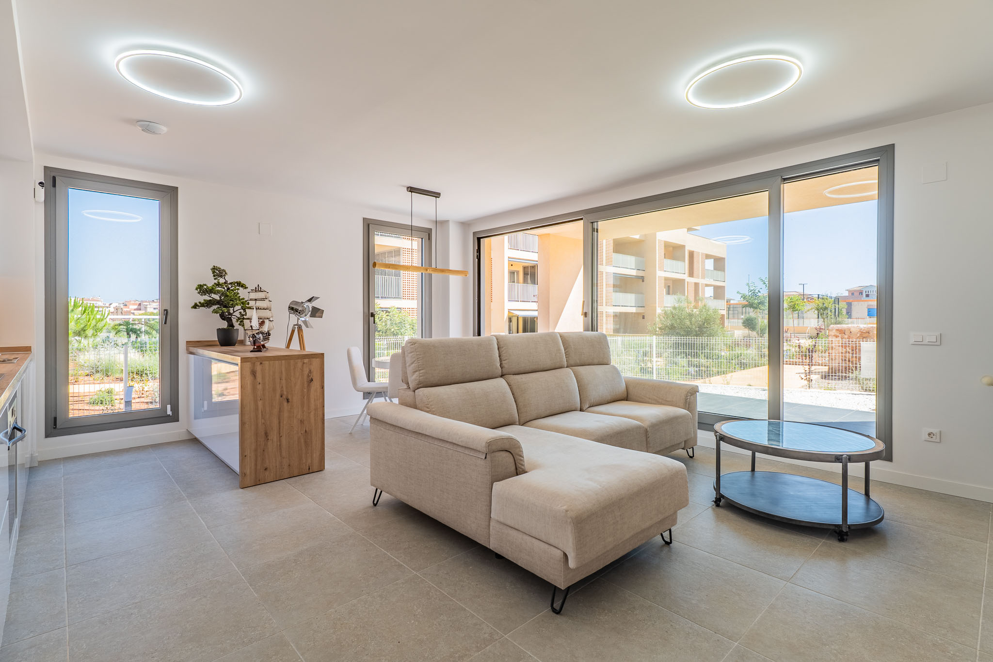Lumineux salon-salle à manger ouvert, avec canapé sectionnel beige et table basse ovale. Grandes baies vitrées offrent lumière naturelle et vues résidentielles. Cuisine ouverte avec comptoir en bois.