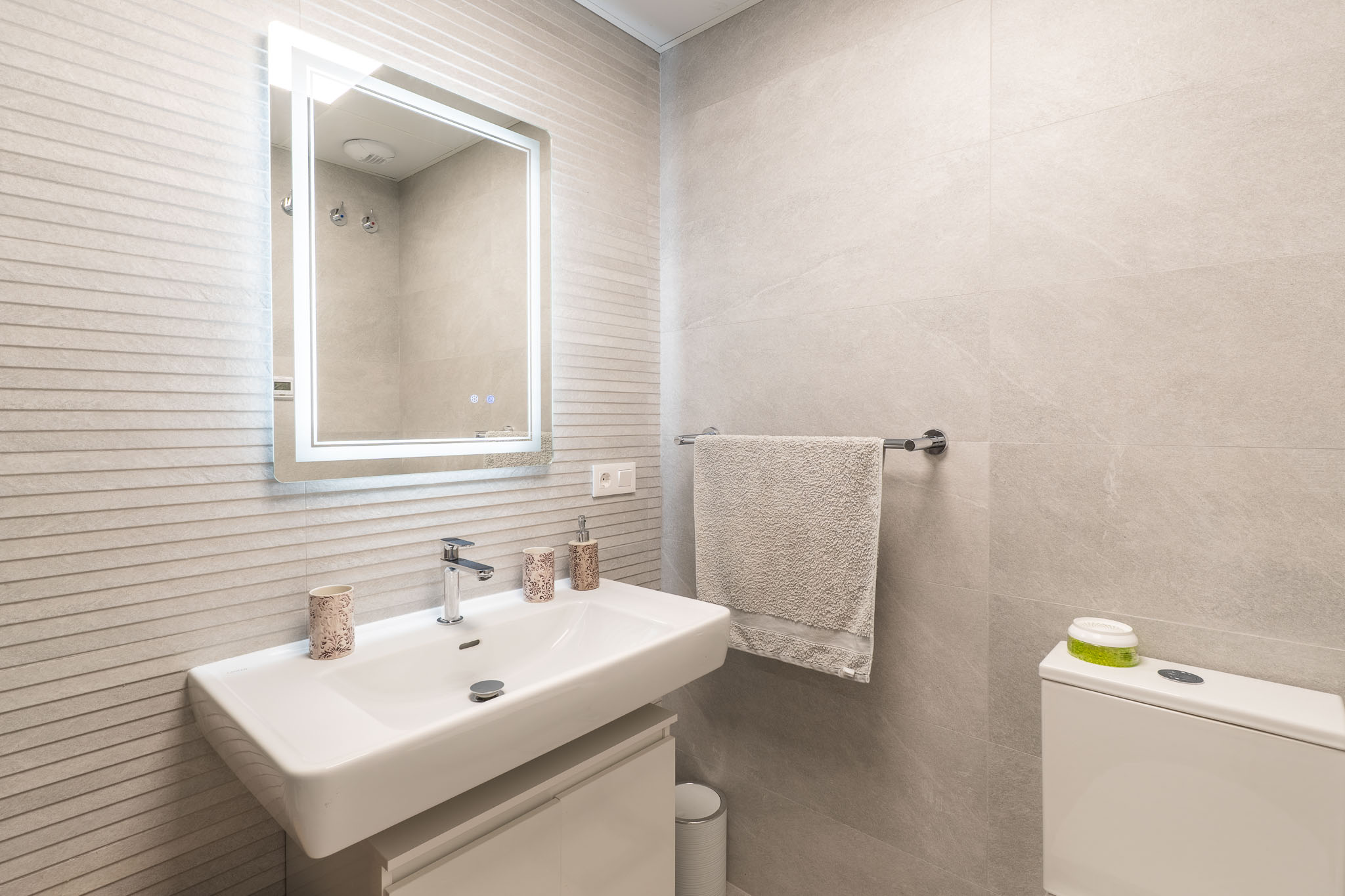 Meuble-lavabo de salle de bain moderne avec lavabo rectangulaire blanc, robinet mural, miroir éclairé et carrelage mural gris texturé.