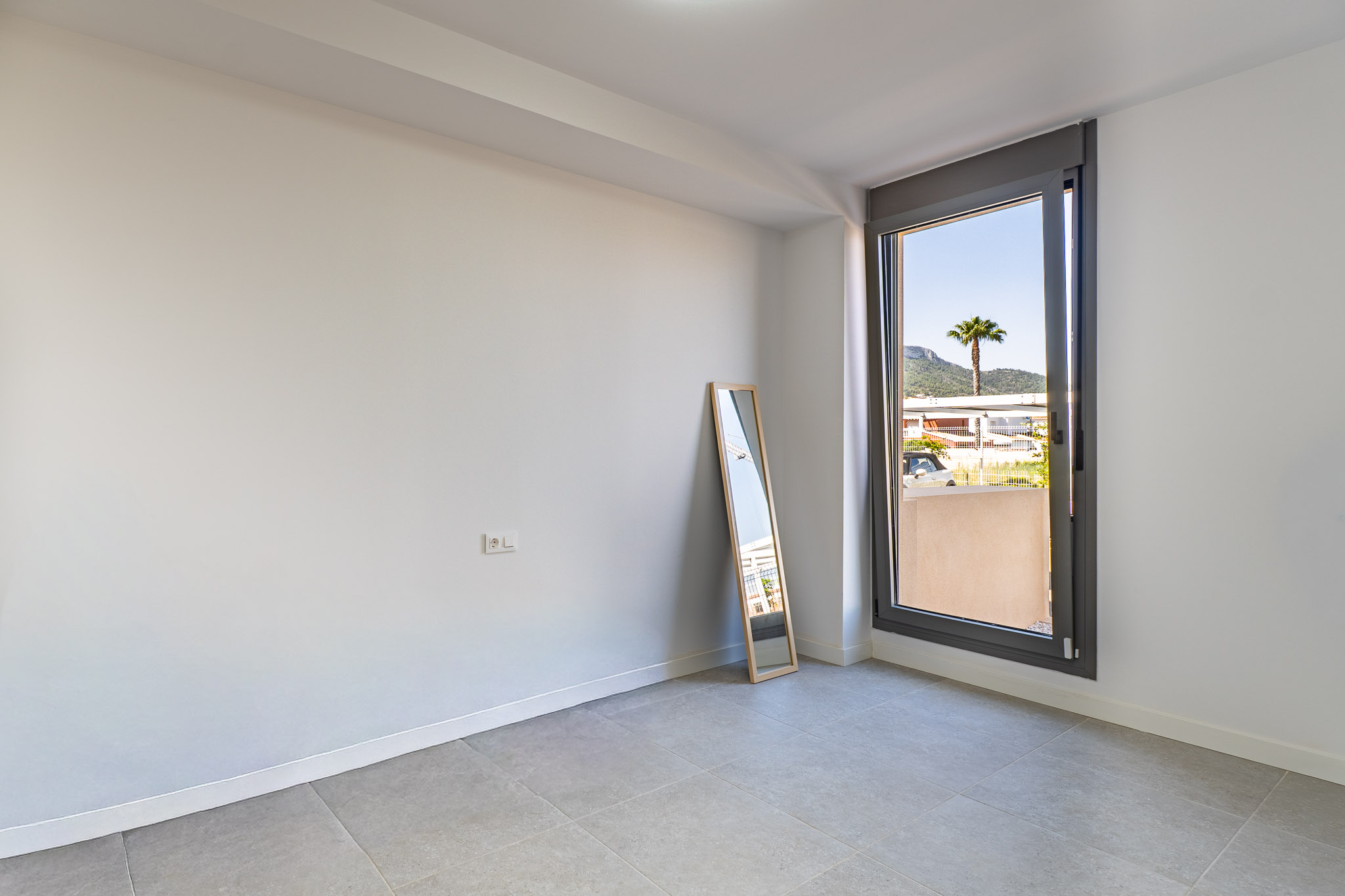 Pièce vide avec murs blancs et carrelage gris, prête à être aménagée. Grande fenêtre offrant une lumière naturelle généreuse et des vues sur un palmier et les montagnes.