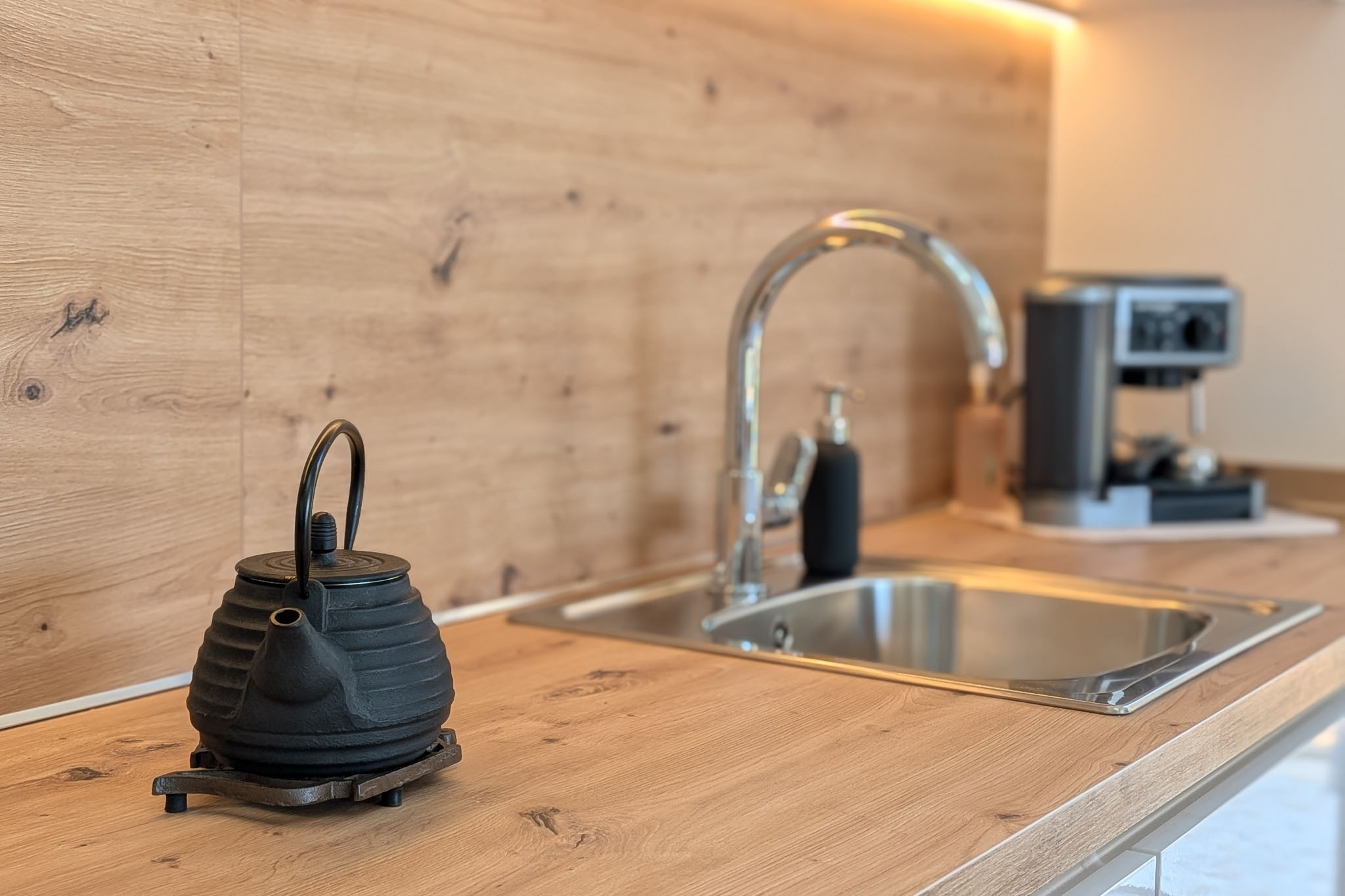 Plan de travail et crédence de cuisine modernes en imitation bois clair. Évier en acier inoxydable avec robinet chromé, éclairage LED sous les meubles et théière en fonte au premier plan.