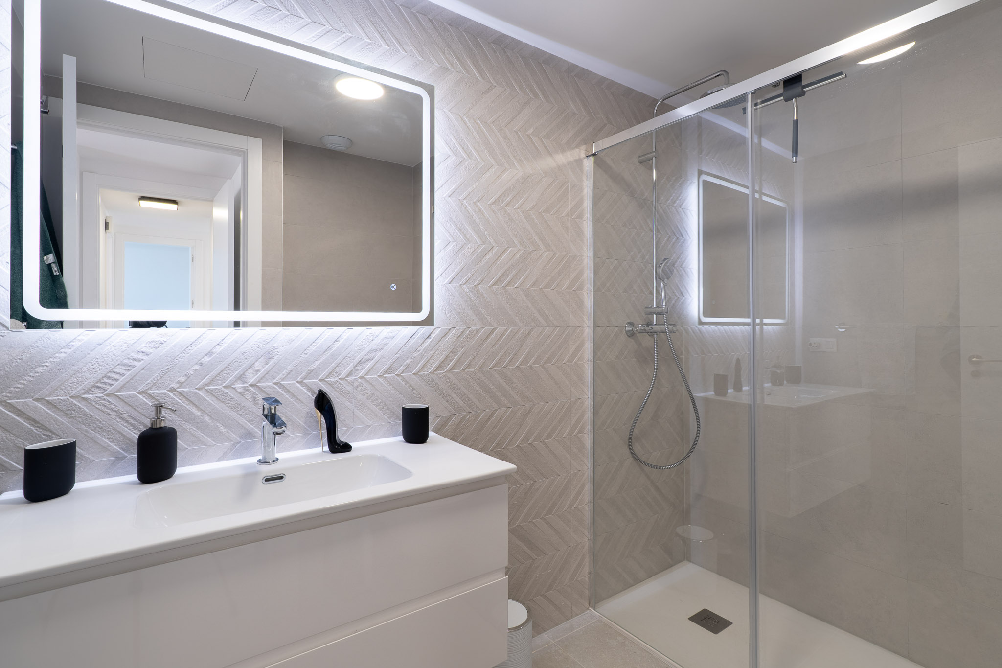 Salle de bain contemporaine avec meuble-lavabo blanc, miroir éclairé et carrelage mural à motif chevrons. La douche vitrée est équipée d'une pomme de douche pluie et de robinetterie chromée.