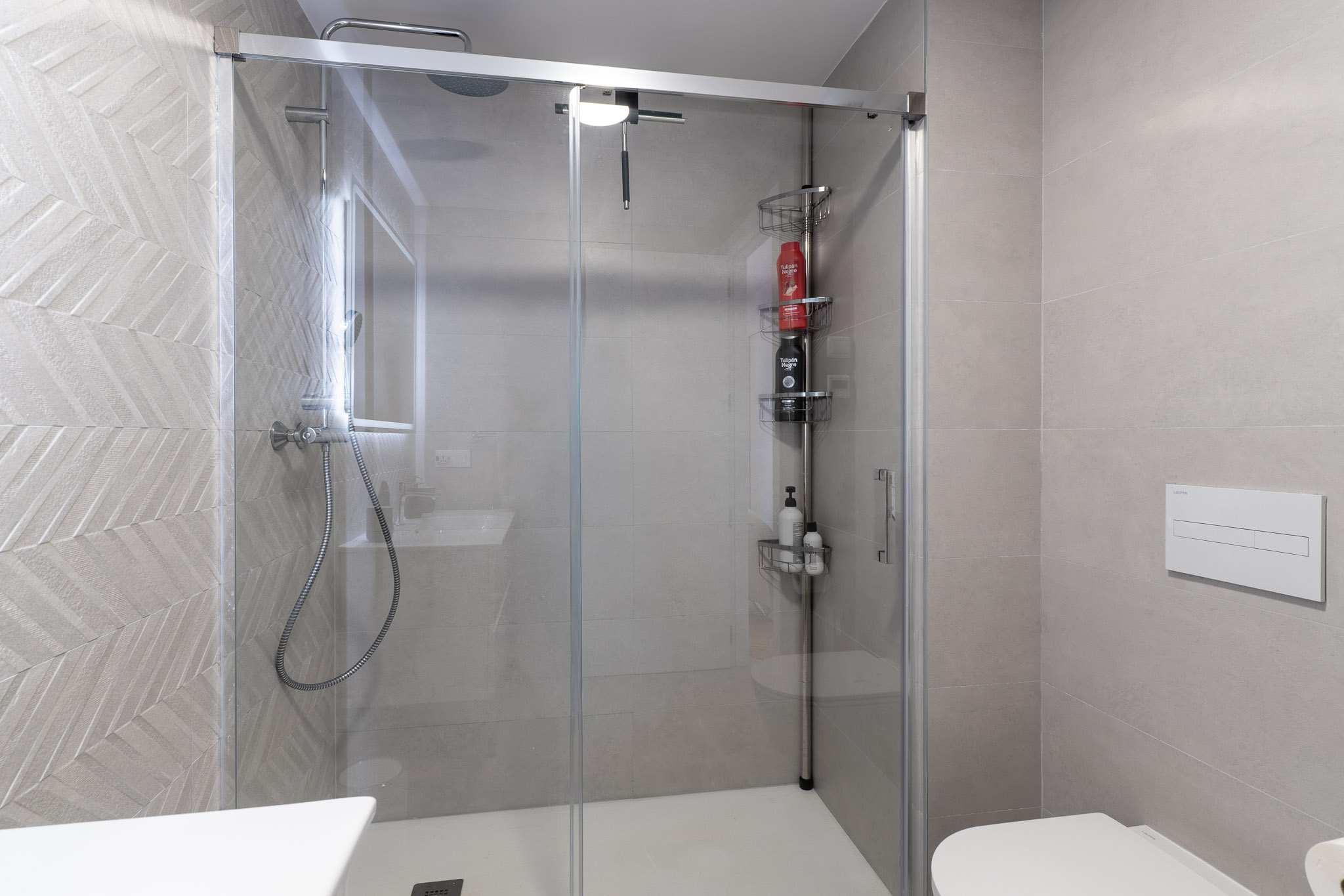 Salle de bain moderne avec cabine de douche vitrée et receveur blanc. Le mur de la douche présente un carrelage gris à motif chevron, complété par une douche à effet pluie et des WC suspendus.