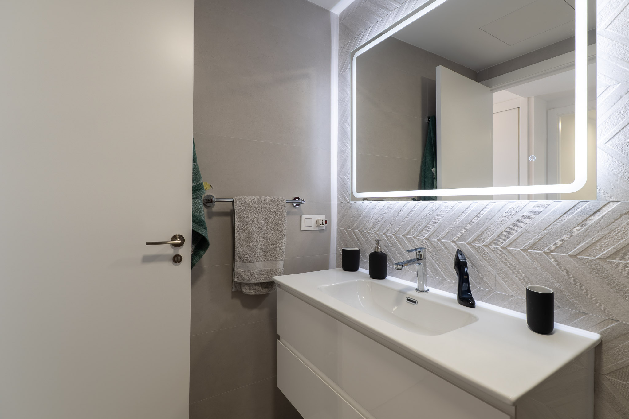 Salle de bain moderne avec meuble-lavabo suspendu blanc, robinetterie chromée et miroir lumineux. Caractérisée par des carreaux muraux texturés à motif chevrons.