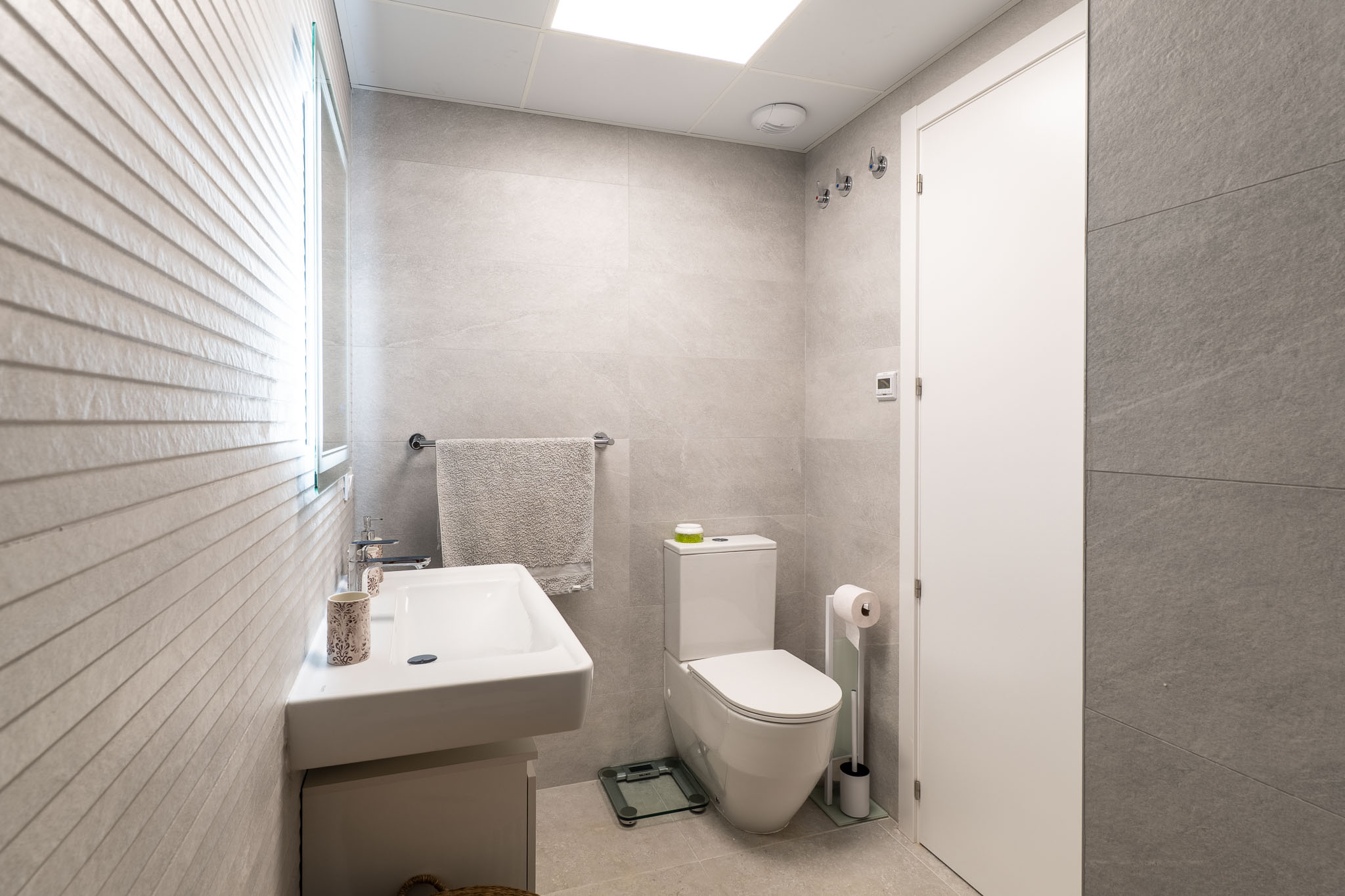 Salle de bain moderne carrelée en gris clair avec lavabo suspendu et toilettes. Un miroir éclairé et un mur texturé apportent du caractère.