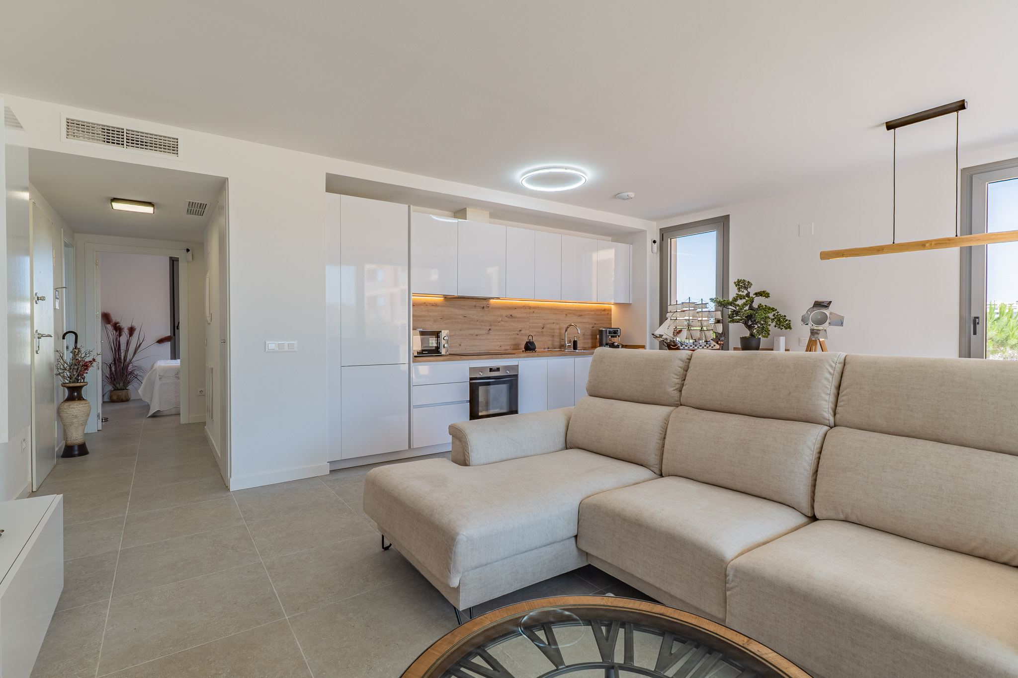 Salon-cuisine ouvert avec canapé sectionnel beige, cuisine moderne aux armoires blanches brillantes et crédence effet bois clair. Carrelage au sol et lumière naturelle.