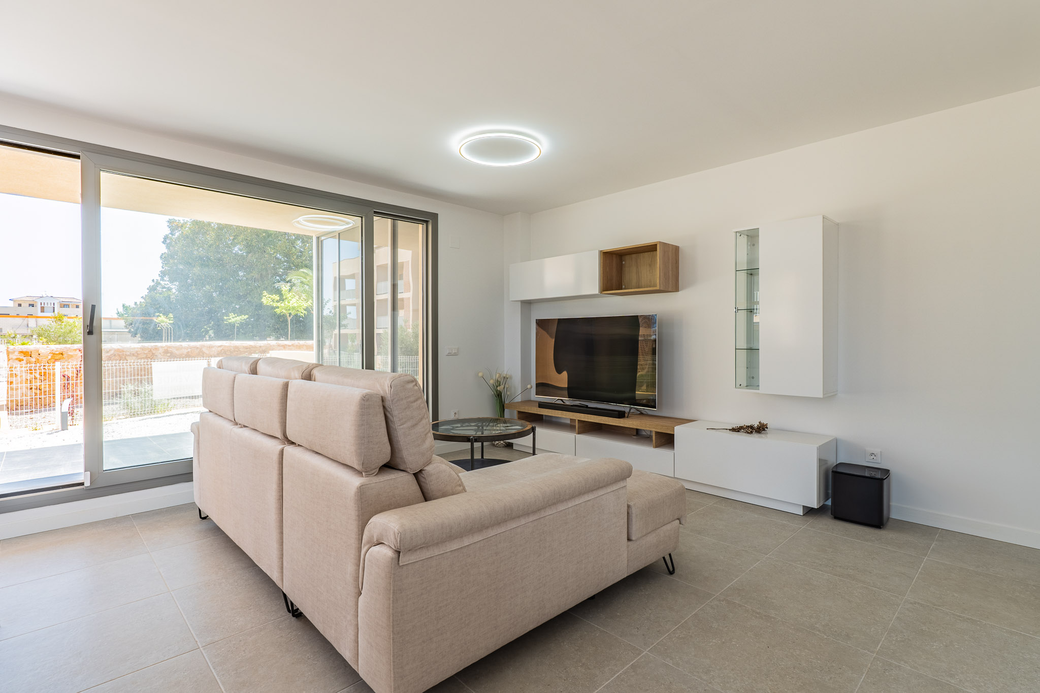 Salon lumineux avec un canapé sectionnel beige, carrelage gris clair et de grandes baies vitrées coulissantes donnant sur l'extérieur. Meuble TV contemporain avec rangements blancs et bois.