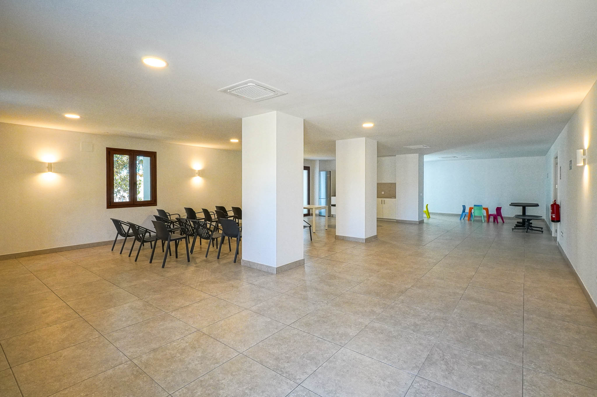 Vaste centre social commun avec carrelage clair, piliers porteurs et éclairage encastré. Comprend un espace de réunion avec chaises et un coin jeux coloré pour enfants.