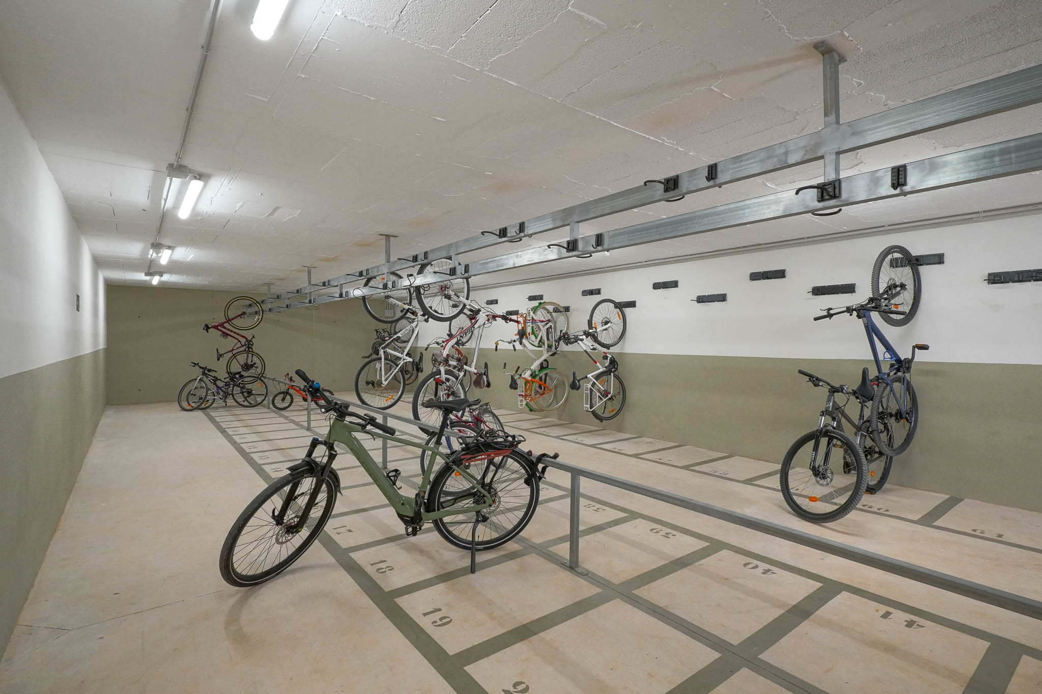 Vaste espace de stationnement commun pour vélos avec emplacements numérotés au sol et supports muraux. Éclairé par des néons, il offre un rangement sécurisé au sein du complexe résidentiel.
