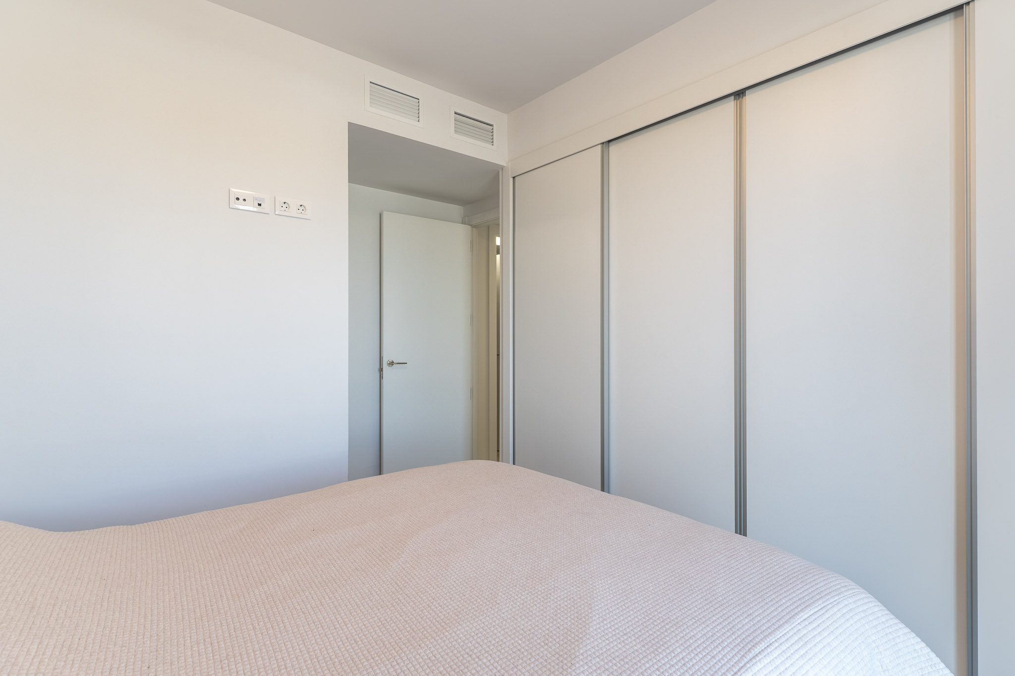 Vue de chambre à coucher avec une grande armoire intégrée à portes coulissantes blanches. Le mur dispose de prises électriques modernes et de grilles de ventilation au-dessus de la porte adjacente.