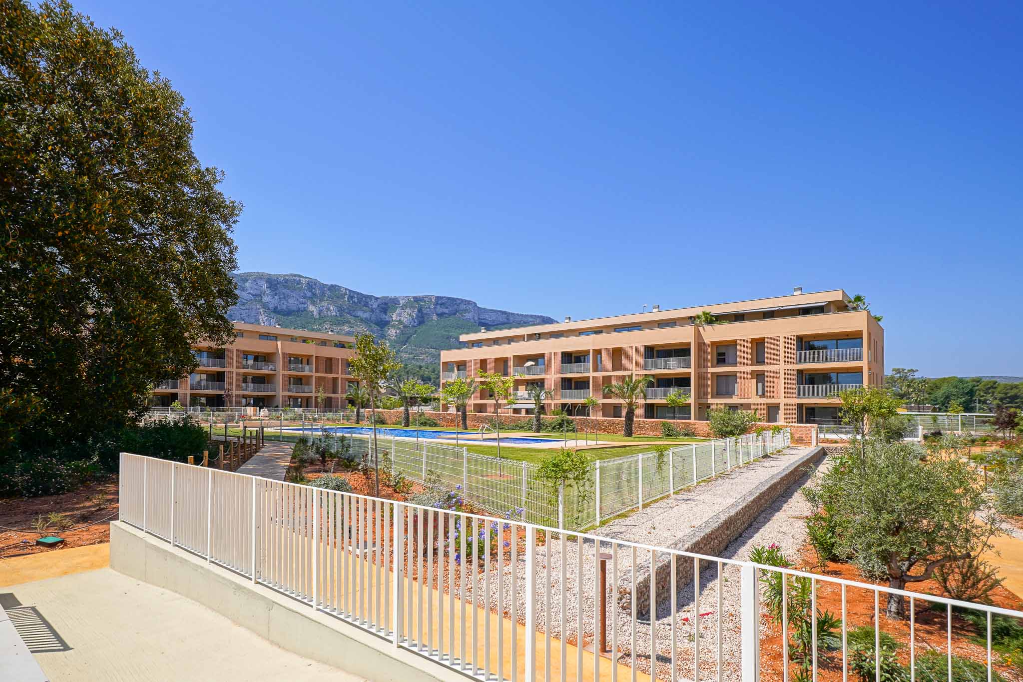 Vue extérieure du complexe résidentiel moderne avec deux bâtiments aux façades claires, une piscine commune et des jardins aménagés. Les montagnes sont visibles en arrière-plan.